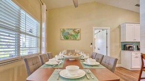 Un comedor con una larga mesa dispuesta para seis personas, una cocina al fondo y un cuadro con temática tropical en la pared.