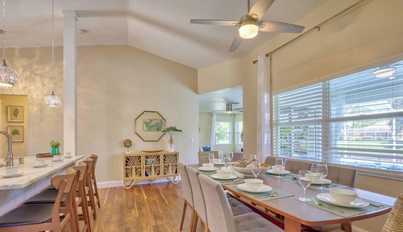 A dining area with a long wooden table set for a meal, a ceiling fan, and a view through large windows.