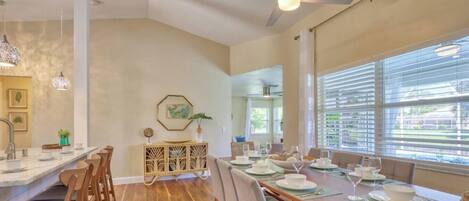 A dining area with a long wooden table set for a meal, a ceiling fan, and a view through large windows.