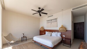Ein Schlafzimmer mit einem Bett, einem Deckenventilator, einer gewobenen Lampe, einem Sessel, einem kleinen Tisch und einer Holzbank.