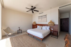 A bedroom with a bed, a ceiling fan, a woven lamp, a chair, a small table, and a wooden bench.
