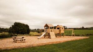 A wooden playground with a slide and swing set, surrounded by a gravel area and a picnic table.