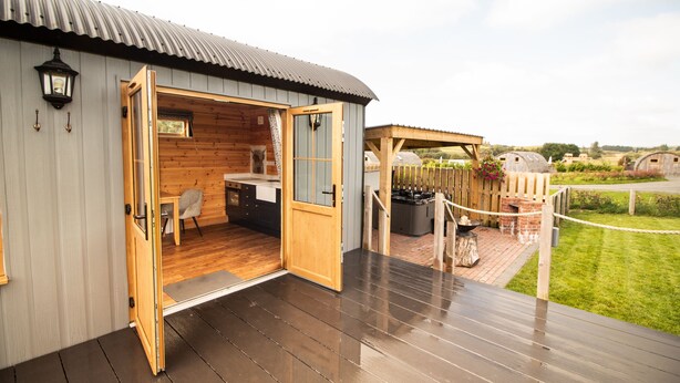 A wooden cabin with an open door leading to a kitchen area, a deck with a barbecue grill, and a fenced garden area.