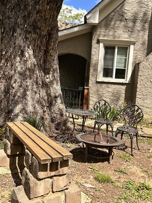 Relax in the serene patio area with a wooden bench and metal table, surrounded by a large tree providing shade.