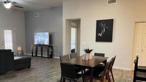 A spacious living room with a dark wood floor, a dining table with chairs, a sofa, and a television on a stand.