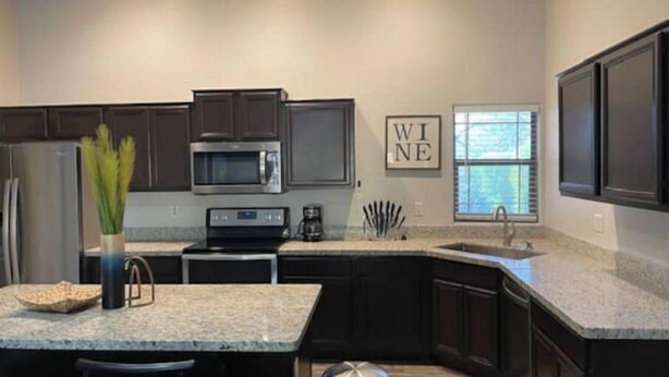 A modern kitchen with dark cabinetry, a granite countertop, stainless steel appliances, and a window with blinds.