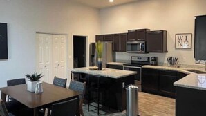 A modern kitchen with dark cabinetry, a stainless steel refrigerator, a microwave, and a coffee maker. There is a dining area with a table and chairs, and a trash can is visible.