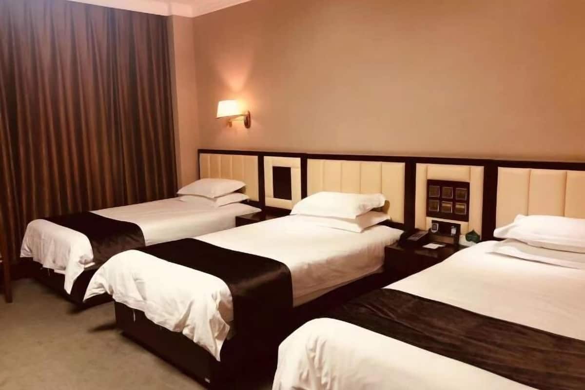 A hotel room with three single beds, a bedside table with a lamp, and a curtain on the left.