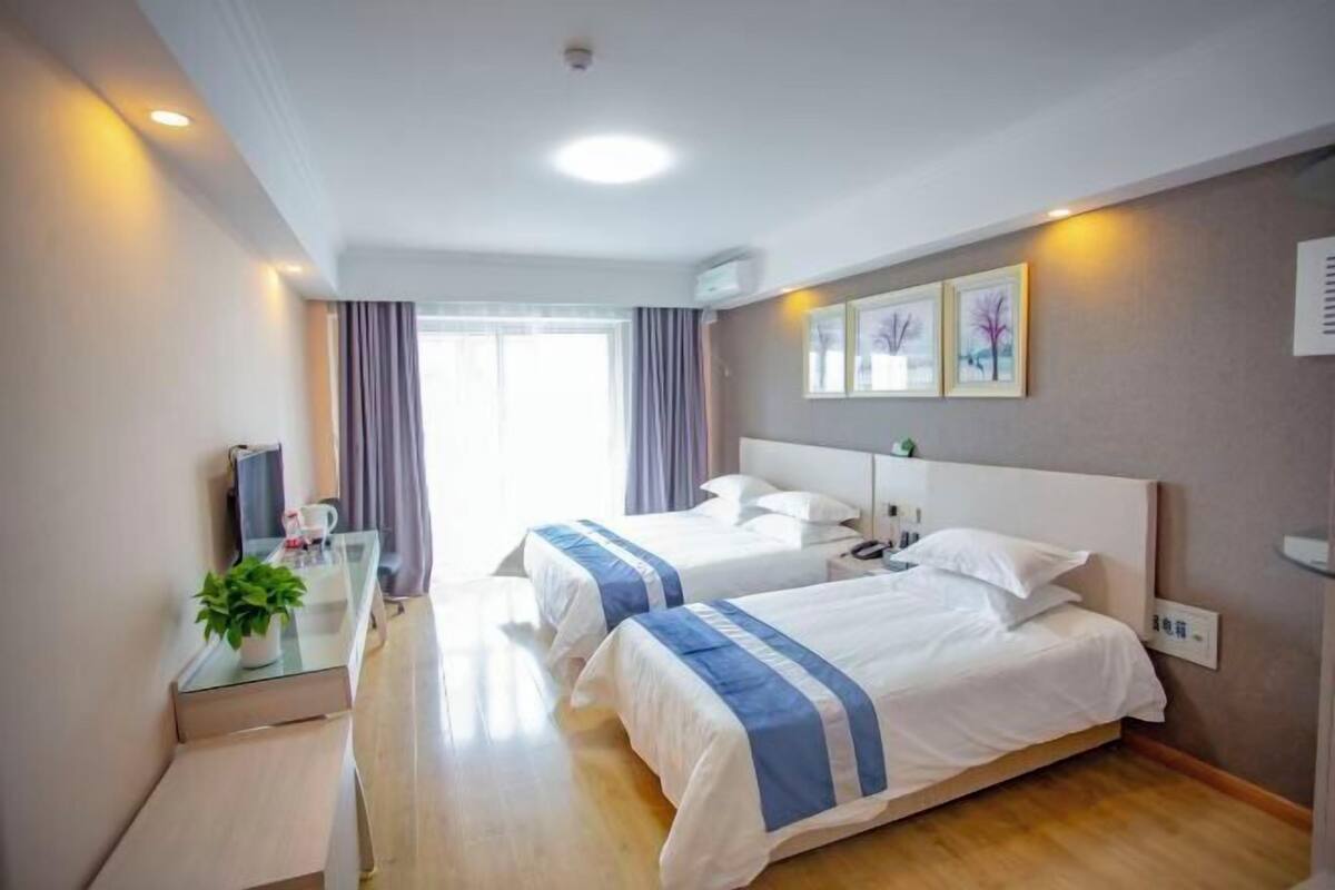 A hotel room with two beds, a glass table with a plant, a bedside table with a lamp, and artwork on the wall.