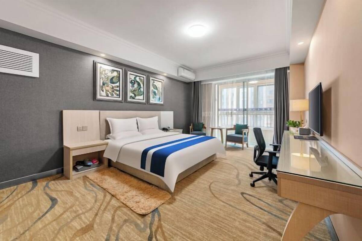 A hotel room with a bed, desk, and chair, a large window, and artwork on the wall.