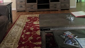 A living room with a large flat-screen TV, wooden entertainment center, and a red patterned rug. - Brown House in the Woods - Barkley Lake Area (Cadiz)