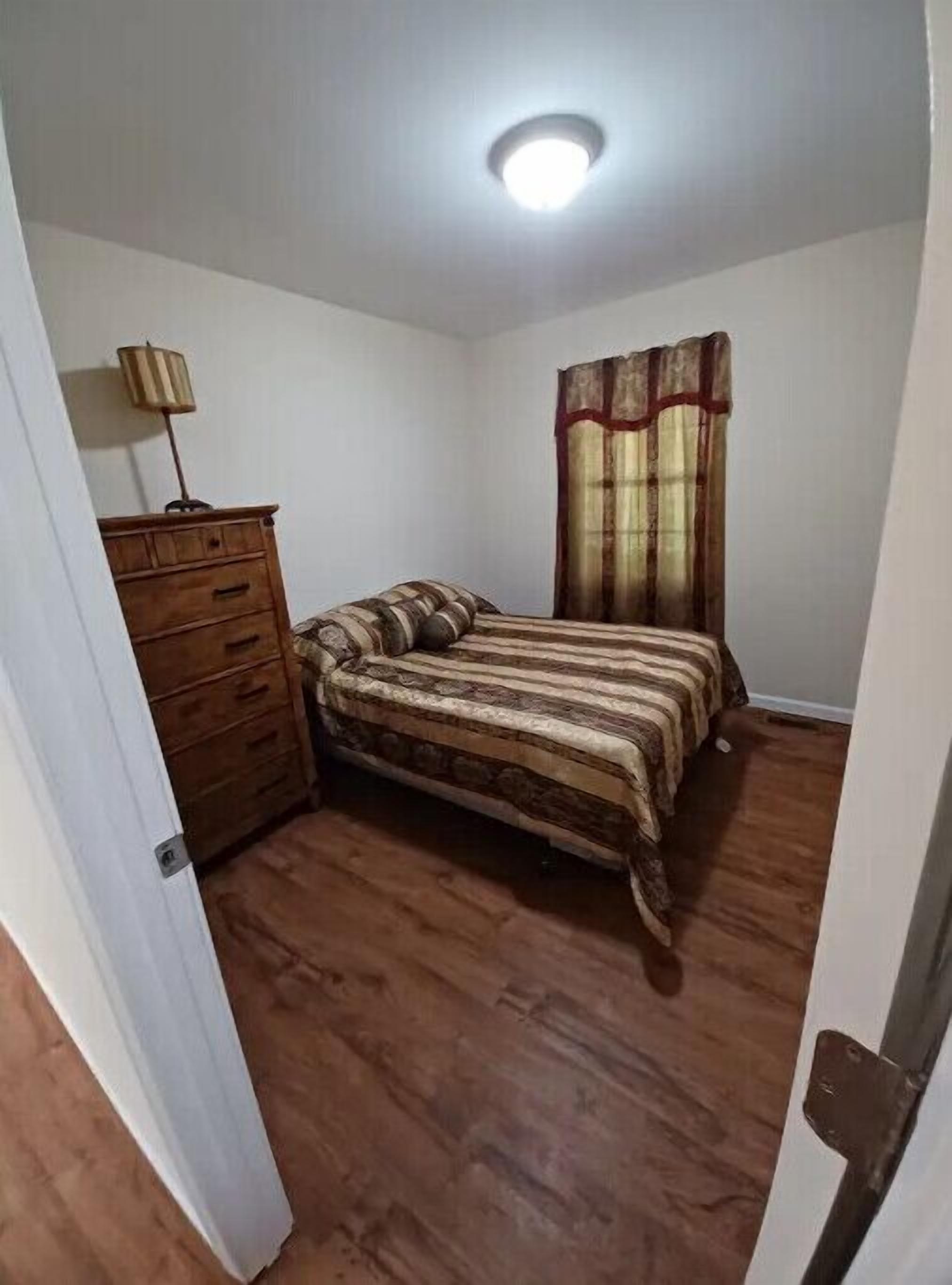 A bedroom with a wooden dresser, a bed with striped bedding, a window with curtains, and a ceiling light.