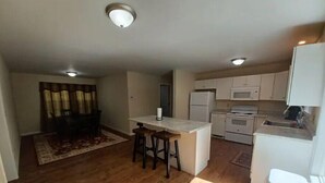 A compact kitchen with a central island, white cabinets, a refrigerator, microwave, and stove. There is a dining area with a table and chairs, and a patterned rug on the floor. - Brown House in the Woods - Barkley Lake Area (Cadiz)