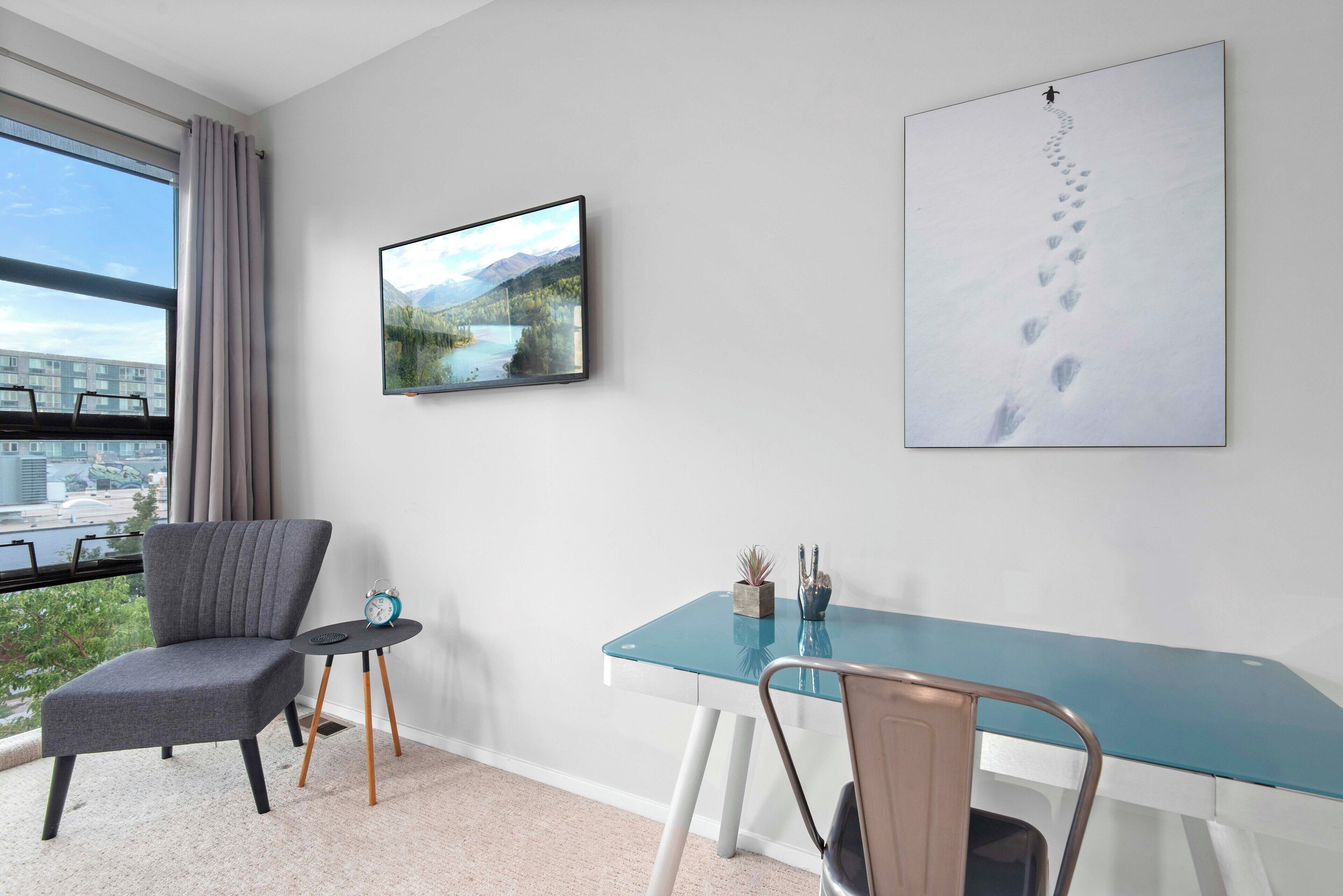 A modern room with a large window, a grey upholstered chair, a glass table with a blue top, and a framed picture of a mountain landscape.