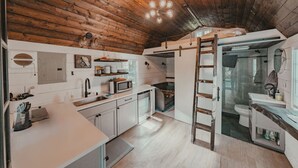 A compact kitchen with white cabinets, a microwave, and a sink. A wooden ladder is leaning against the wall. - Clay Baby- luxury shed conversion on the river + Epic Sauna + Cold Plunge (Sultan)