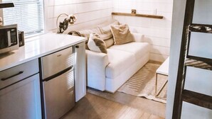 A compact kitchen with a microwave, a sink, and a white countertop. There is a white sofa with a blanket and pillows, and a wooden floor. - Clay Baby- luxury shed conversion on the river + Epic Sauna + Cold Plunge (Sultan)