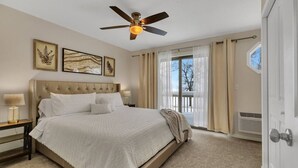 A bedroom with a bed, a ceiling fan, a nightstand, a window with curtains, and a door.