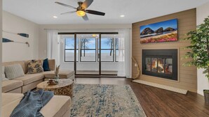 A living room with a sofa, coffee table, fireplace, ceiling fan, and a large window with a view.