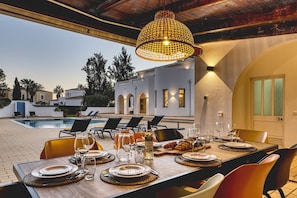 An outdoor dining area with a pool, lounge chairs, and a pergola with a hanging light fixture. - Villa Beleza - Elegant Villa on Penina Golf Course (Alvor)