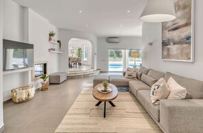 A modern living room with a large sofa, a coffee table, and a TV. There is a fireplace, a dining area, and a view of a pool. - Villa Beleza - Elegant Villa on Penina Golf Course (Alvor)