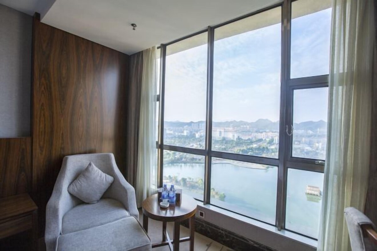 A room with a large window, a chair, a small table with bottles, and a view of a cityscape and river.