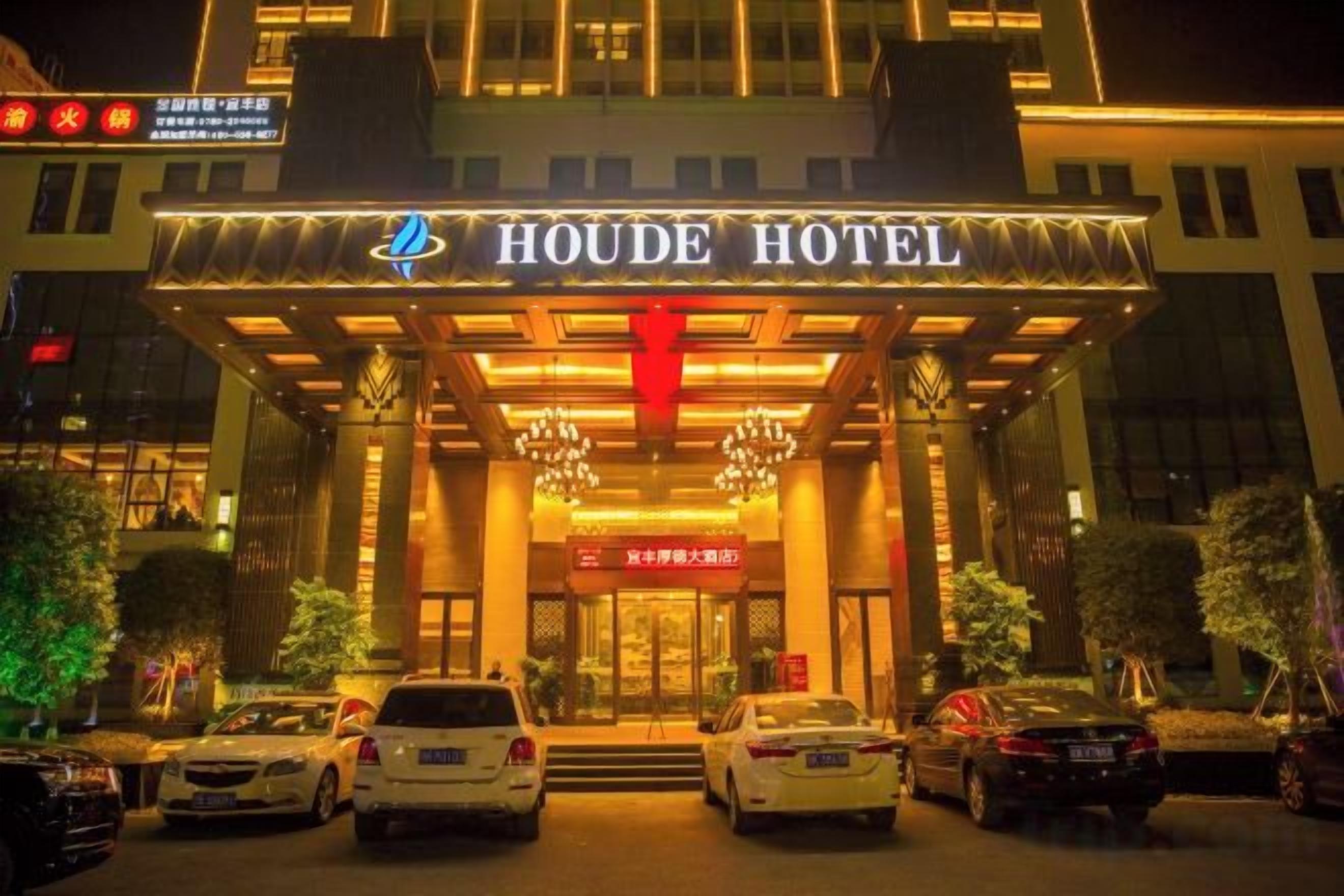 The exterior of the Houde Hotel with a prominent entrance, a red banner, and several cars parked in front.