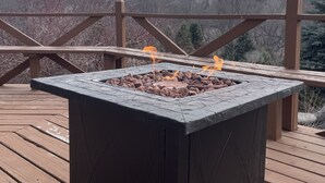 Et firkantet udendørs bålsted på en træterrasse med udsigt til et skovområde.