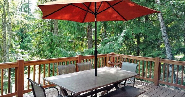 Une terrasse en bois, agrémentée d’une table et de chaises, entourée d’une balustrade en bois, le tout se fondant dans un décor de feuillus verdoyants.