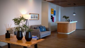 A modern interior with a wooden reception desk, a grey sofa with blue cushions, a wooden dining table with black vases and books, and a wall-mounted lamp. - Pero de teive Bay Apartments Hotel (Ponta Delgada)