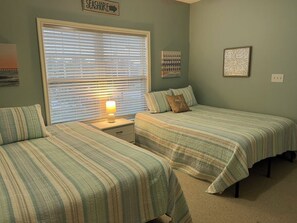 A bedroom with two beds, a nightstand with a lamp, a window with blinds, and wall art. - Relax in Lewes Condo near the beaches! (Lewes)
