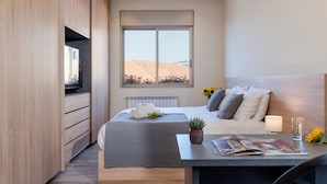 Quarto moderno com uma cama grande, uma mesa de cabeceira com abajur, uma pequena planta em vaso e uma janela com vista para edifícios e girassóis.