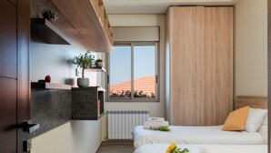 Une chambre d’hôtel moderne dotée d’une grande fenêtre, d’un lit avec une literie blanche, d’une tête de lit en bois et d’une étagère intégrée agrémentée de plantes.