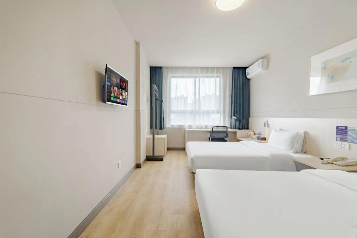 Una camera d'albergo con due letti, una scrivania, una sedia, una finestra con tende, un televisore a parete e un condizionatore d'aria.
