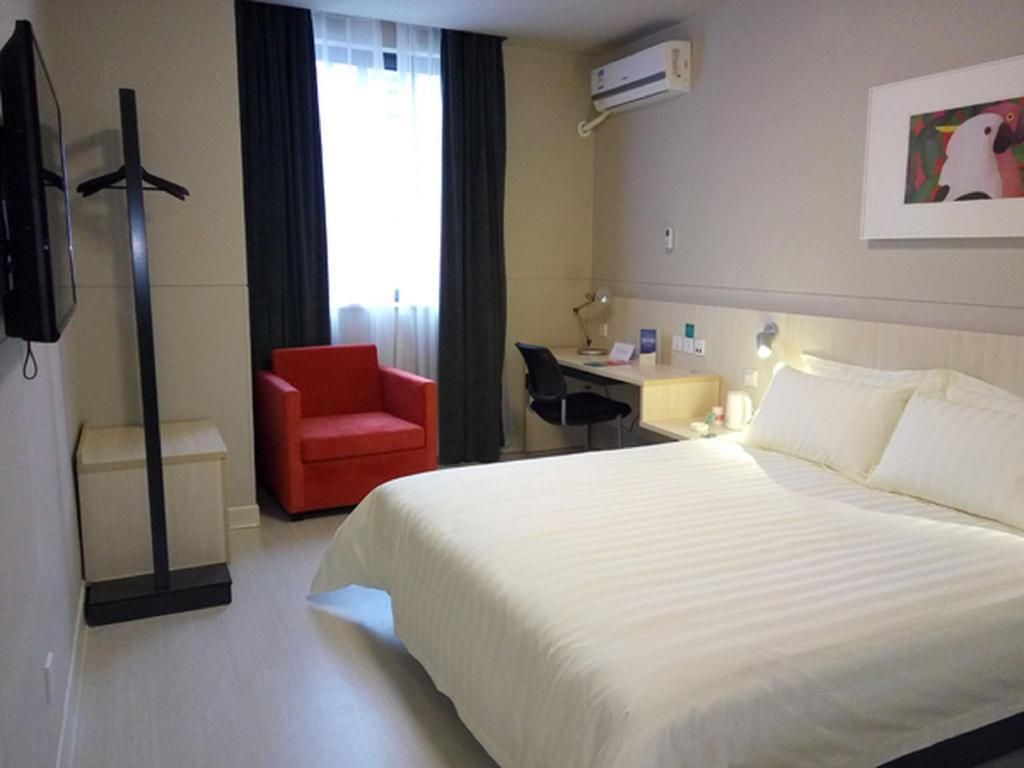 A hotel room with a bed, a desk, a chair, a coat rack, and a television.
