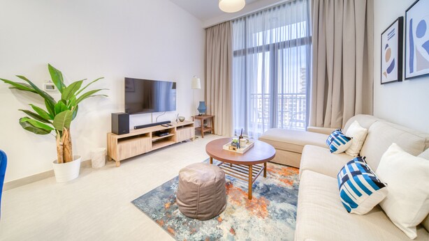 A modern living room with a flat-screen TV, a sofa, a coffee table, and a potted plant. - Sophisticated 1BR at Town Square Rawda by Deluxe Holiday Homes (Dubai)