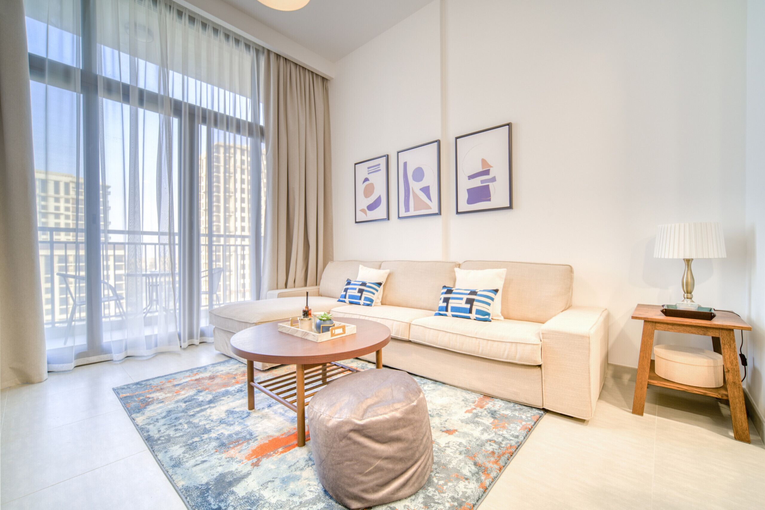A modern living room with a sofa, ottoman, coffee table, and a lamp. There are large windows with curtains, a city view, and three framed pictures on the wall.