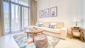 A modern living room with a sofa, ottoman, coffee table, and a lamp. There are large windows with curtains, a city view, and three framed pictures on the wall. - Sophisticated 1BR at Town Square Rawda by Deluxe Holiday Homes (Dubai)