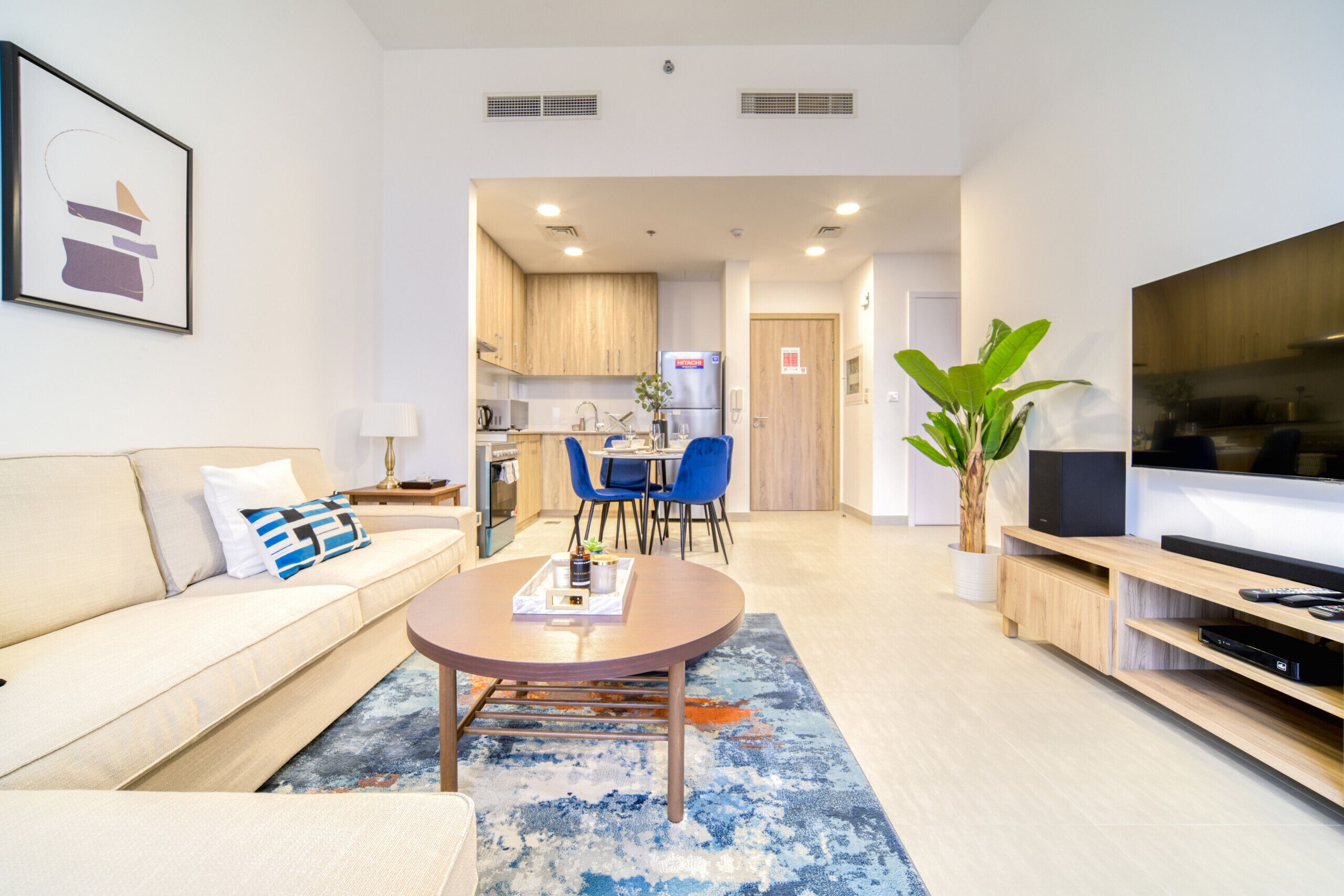 A modern living room with a sofa, coffee table, and TV stand. There is a dining area with chairs and a table. The kitchen is visible in the background.