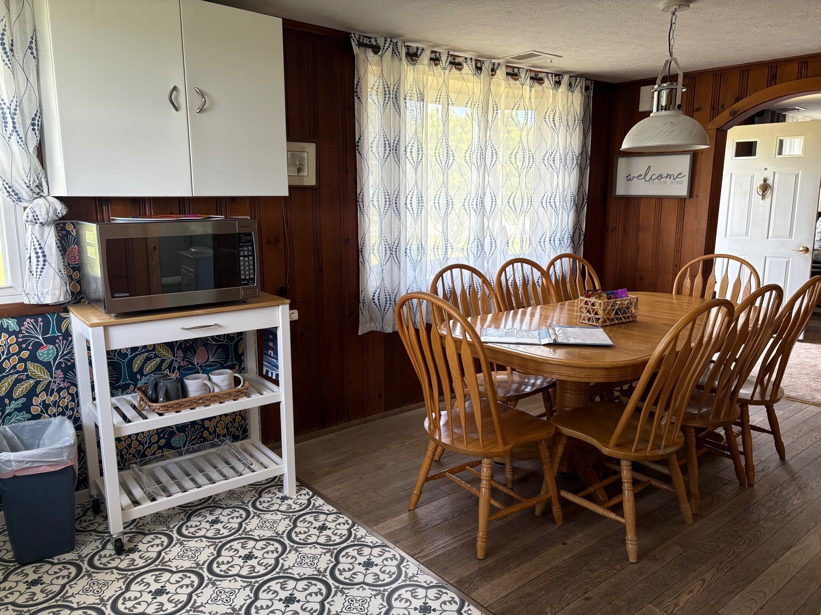 A dining area with a wooden table and chairs, a white cabinet, a microwave on a small shelf, patterned curtains, and a welcome sign.