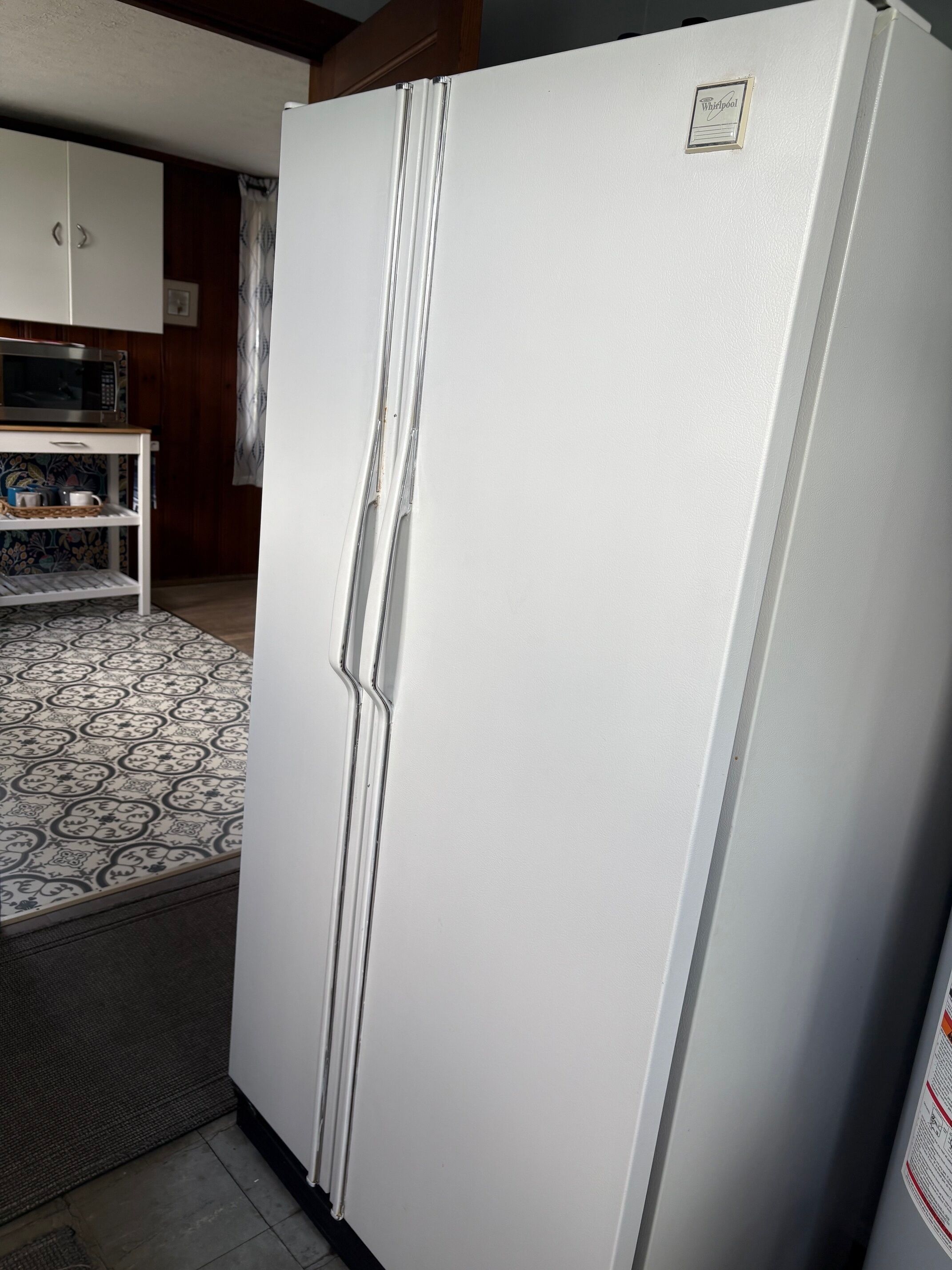 A white refrigerator with a freezer compartment, a microwave above it, and a patterned rug on the floor.