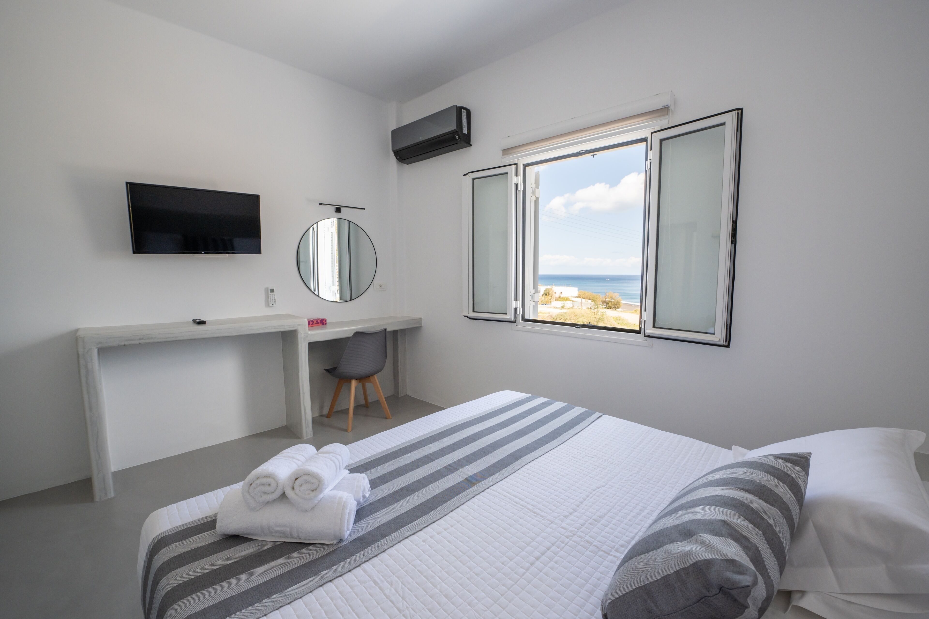 A hotel room with a bed, a desk with a chair, a TV, a window with a beach view, and a mirror.