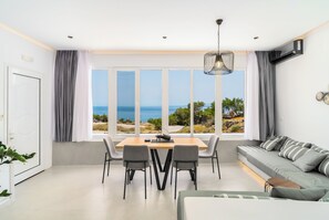 Un salon moderne doté d’une grande fenêtre donnant sur la mer, d’un coin repas avec une table et des chaises, et d’un canapé agrémenté de coussins à rayures.