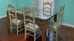 A dining area with a glass-top table surrounded by four chairs, a wooden wall clock, and a window with blinds. - Seaside Retreat (Wachapreague)