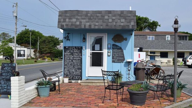 A small, blue building with a white door, a signboard with various texts, and outdoor seating with chairs and potted plants. - Seaside Retreat (Wachapreague)