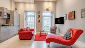 Una sala moderna con un sofá rojo, un sillón rojo y una mesa de centro de madera. Hay una zona de cocina con microondas y armarios.