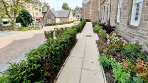 Una calle estrecha flanqueada por edificios de piedra, con aceras pavimentadas y setos y macizos de flores bien cuidados.