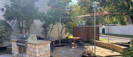 A backyard with a stone barbecue, a wooden table and chairs, and a fire pit.