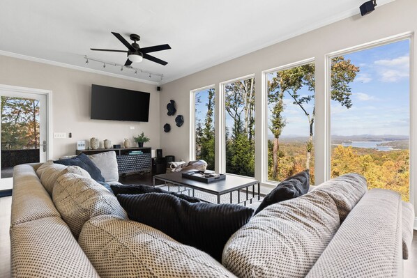 A modern living room with a large sofa, a coffee table, and a flat-screen TV mounted on the wall. There are large windows offering a view of trees and a lake. - Modern Mountain Oasis • 5BR with Million-Dollar Lake Views • EV• Hot Tub • PetOK (Blue Ridge)