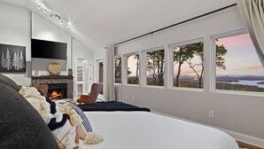 A bedroom with a large bed, a fireplace, a TV, and a view of a sunset over a landscape. - Modern Mountain Oasis • 5BR with Million-Dollar Lake Views • EV• Hot Tub • PetOK (Blue Ridge)