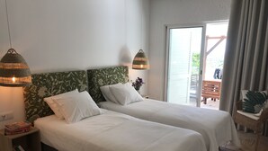 Quarto de hotel com duas camas, mesa de cabeceira com livros, uma cadeira de vime e vista para o exterior.